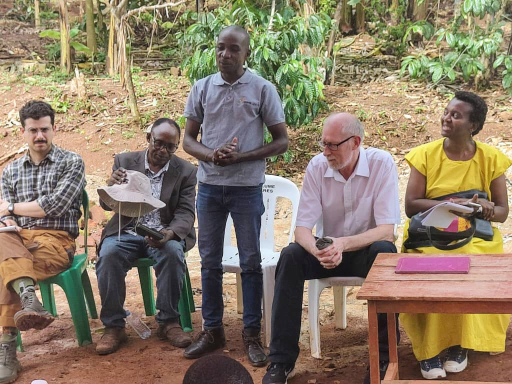 The image shows a group of people seated around a table, some engaged in conversation or presentation, with natural surroundings and a mix of casual and formal attire.AI-generated content may be incorrect.