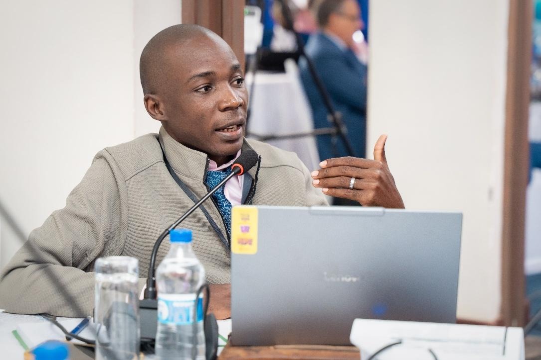 The image shows a person in a suit and tie, sitting at a table with a laptop and a water bottle, engaged in a discussion, with other people and chairs in the background.AI-generated content may be incorrect.