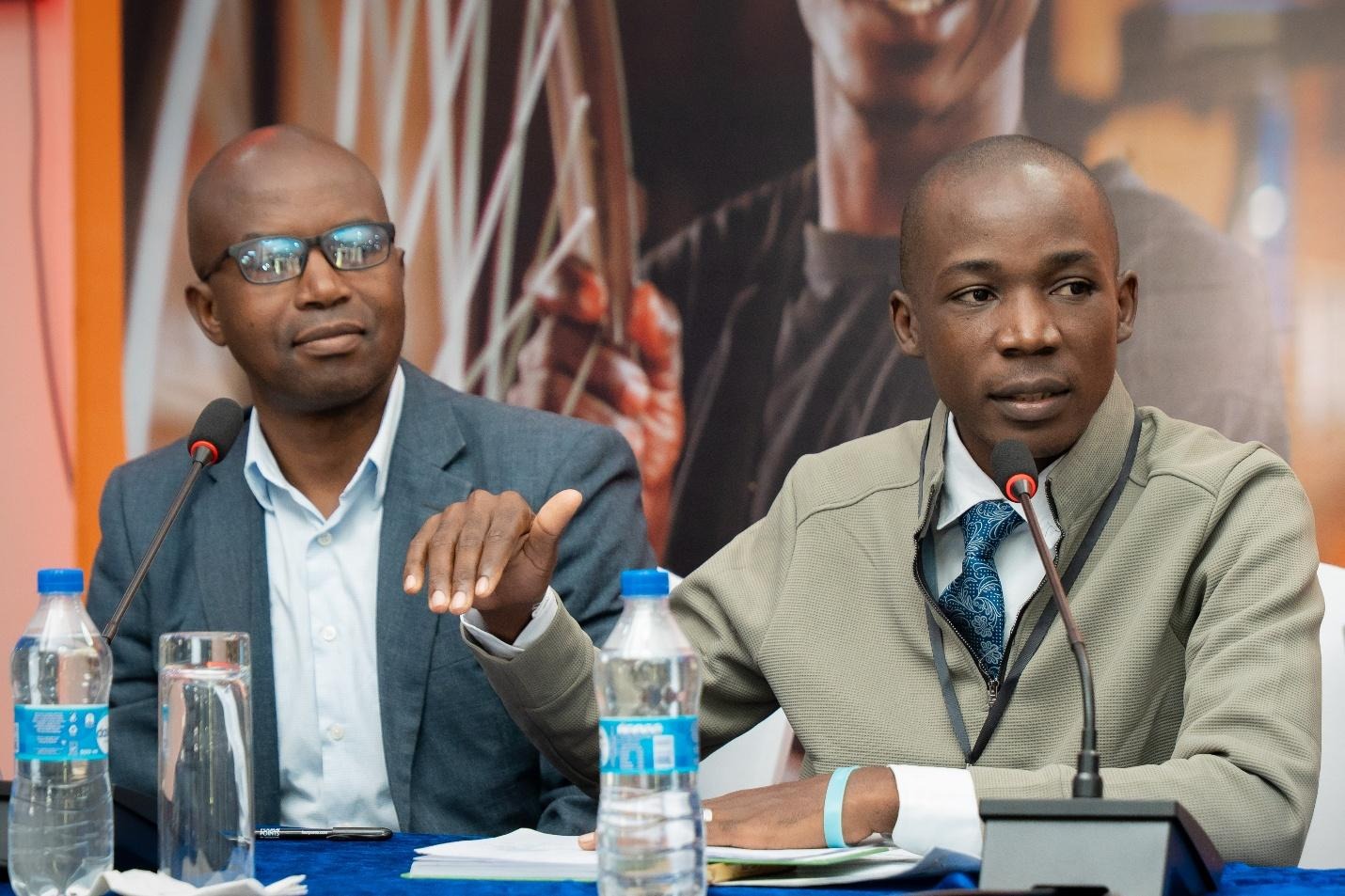 The image shows two individuals in a meeting setting, seated at a table with water bottles, a laptop, and a tablet, engaged in a discussion with a focus on points being made.AI-generated content may be incorrect.