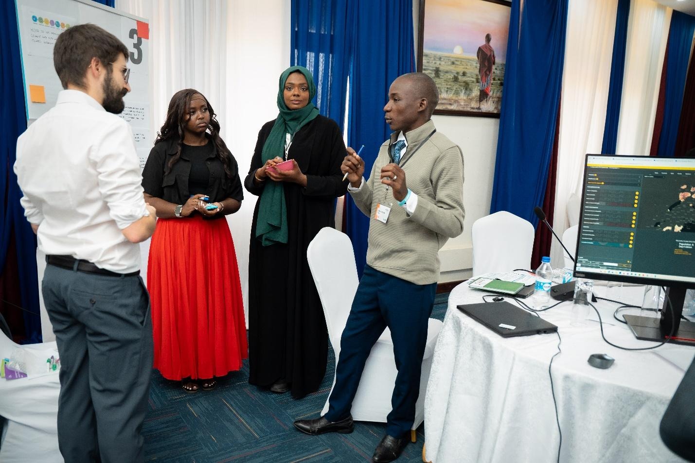 The image depicts a group of individuals in a professional setting, likely a conference or meeting, engaging with technology, with one person speaking into a device, and a backdrop featuring a blue wall and a wallpaper with a large image or design.AI-generated content may be incorrect.