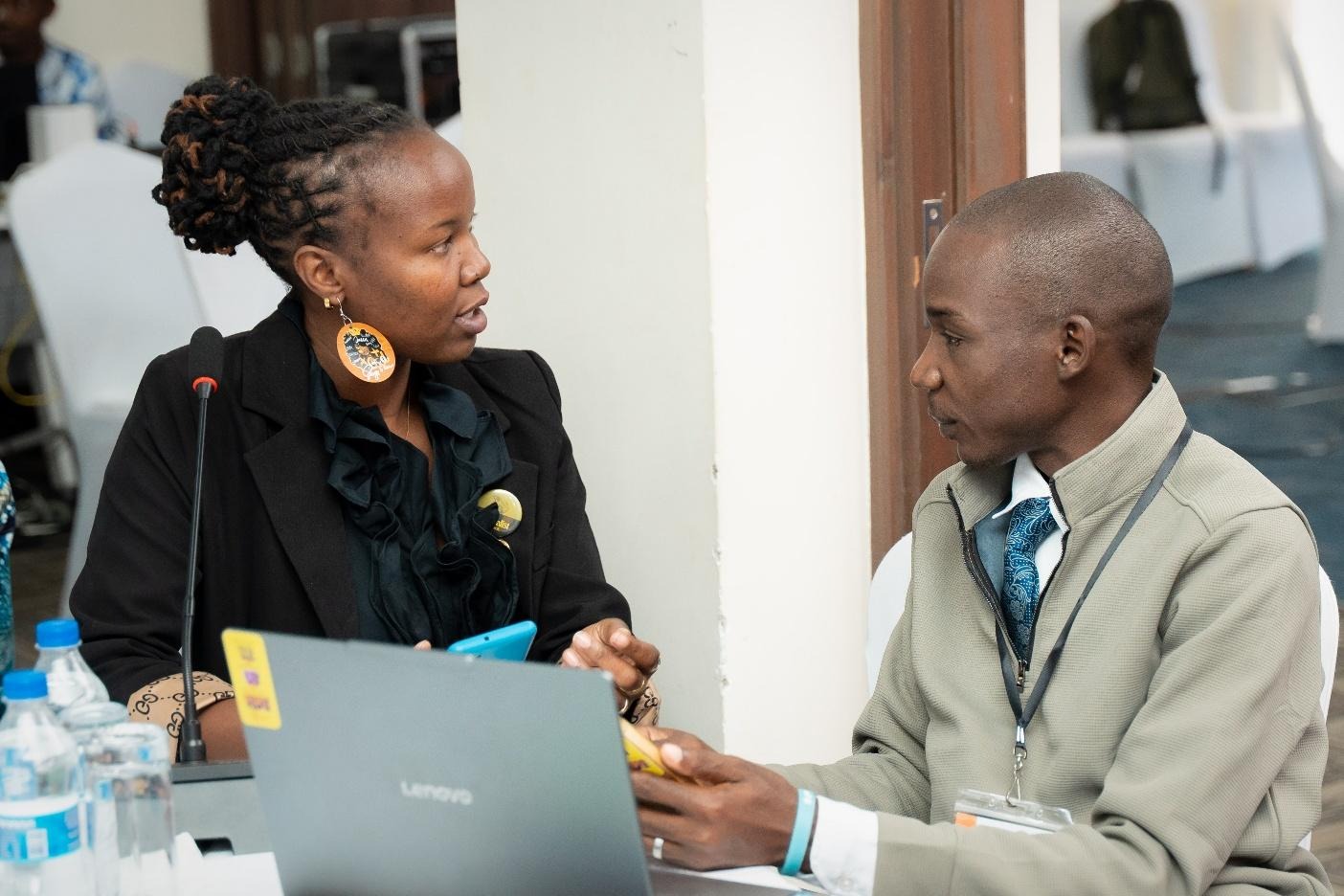 The image shows two people engaged in a conversation at a table, with a laptop and various items on the surface, in an indoor setting with a blurred background.AI-generated content may be incorrect.