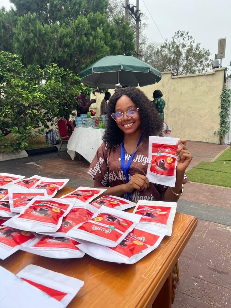 The image shows a person holding a stack of red envelopes, possibly for a fundraising or promotional event, with a clear view of greenery and a white tent in the background.AI-generated content may be incorrect.