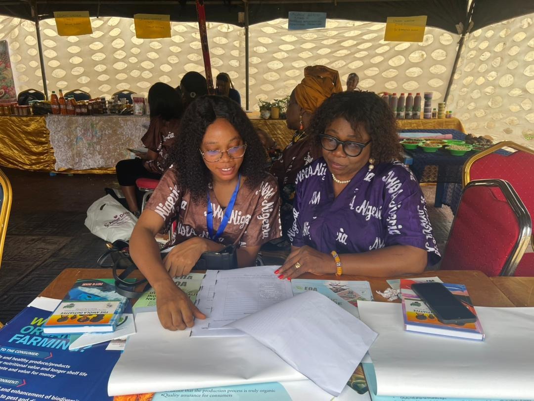 The image shows two people seated at a table with papers and documents, likely engaged in a discussion or presentation about food quality, safety, and nutrition, with a focus on organic produce and pest control.AI-generated content may be incorrect.