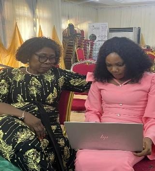 The image shows two individuals, one in a black and gold patterned dress and another in a pink dress, sitting in front of a red carpet and a laptop, possibly in an office setting.AI-generated content may be incorrect.