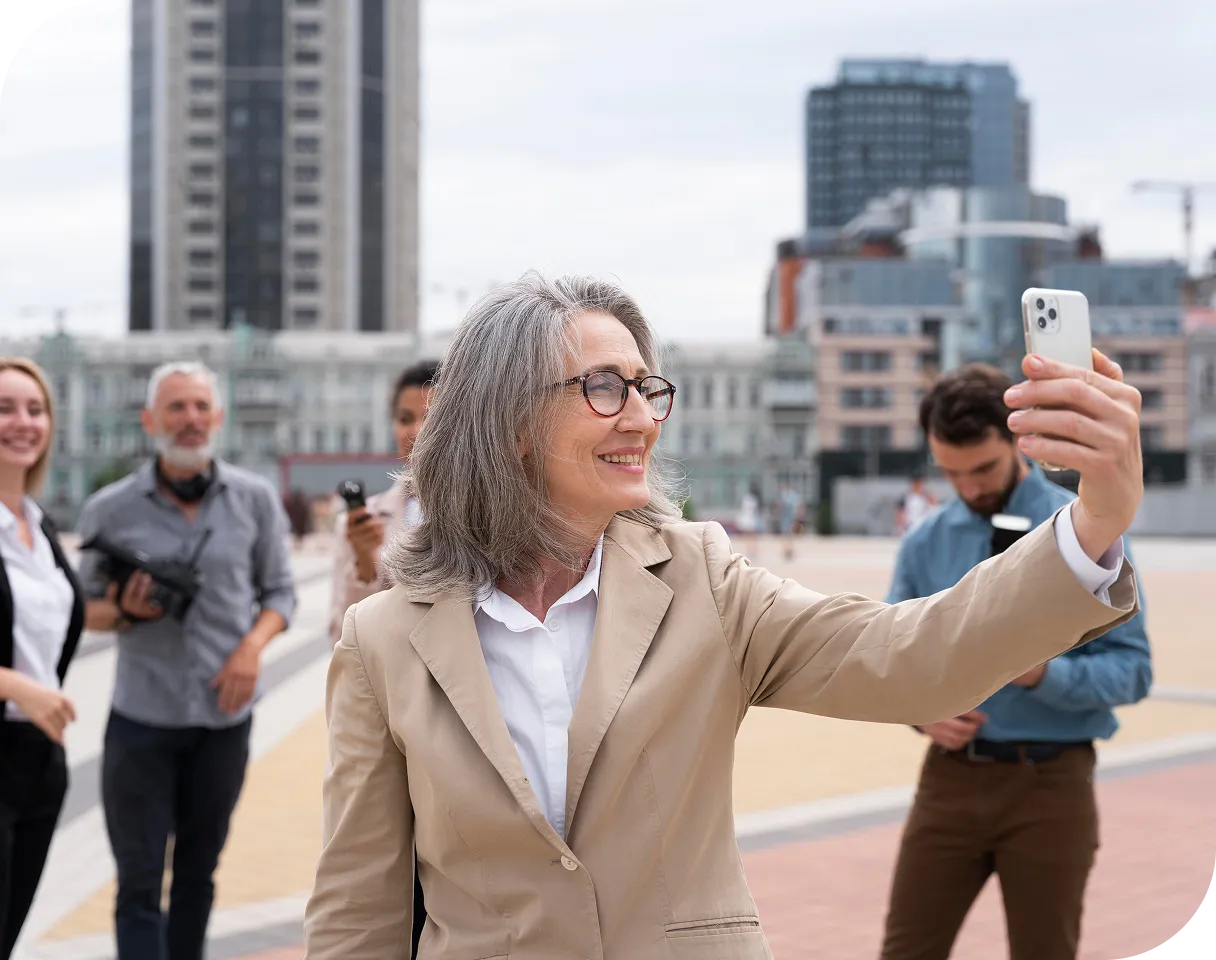 Eine lächelnde Frau mittleren Alters mit Brille macht draußen ein Selfie, hinter ihr sind Menschen und Gebäude zu sehen.