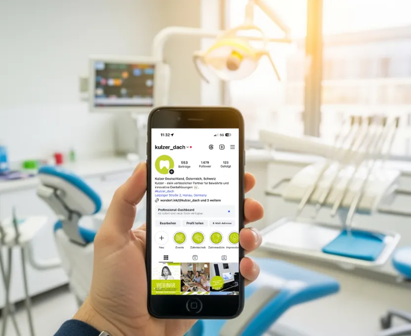 Eine Hand, die ein Smartphone hält, auf dem das Instagram-Profil von Kulzer Dental zu sehen ist, in einer hellen Zahnarztpraxis.