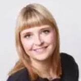Eine lächelnde Frau mit blonden Haaren und Pony, die ein schwarzes Oberteil trägt, vor einem einfarbigen Hintergrund.