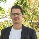 Ein Mann mit Brille und schwarzem Blazer steht im Freien, im Hintergrund sind Bäume zu sehen.