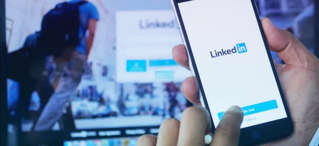 Eine Person hält ein Smartphone in der Hand, auf dessen Bildschirm der Anmeldebildschirm der LinkedIn-App zu sehen ist, während im Hintergrund die LinkedIn-Website unscharf im Hintergrund zu erkennen ist.
