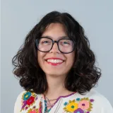 Eine lächelnde Frau mit lockigem braunem Haar, Brille und einem bunten Oberteil mit Blumenmuster vor einem schlichten hellen Hintergrund.