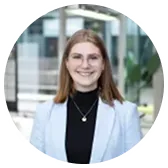 Eine lächelnde junge Frau mit schulterlangem Haar, die in einem hellen Blazer und einem schwarzen Oberteil steht.