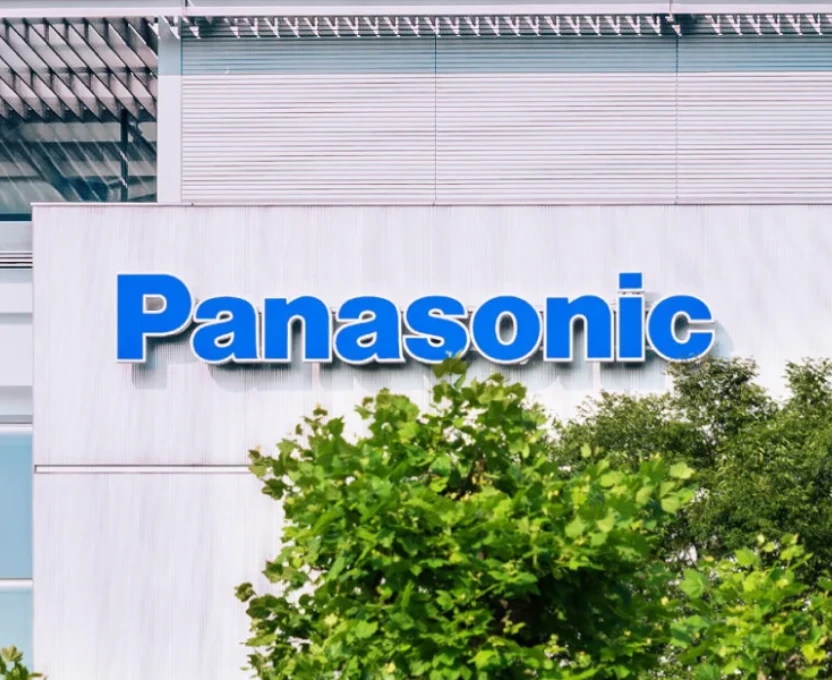 Das Panasonic-Logo in fetten blauen Buchstaben auf einer weißen Gebäudefassade über grünen, blättrigen Bäumen.