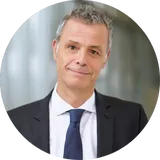 Ein Mann in Geschäftskleidung mit kurzen grauen Haaren, der vor einem unscharfen Hintergrund sanft lächelt.