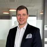 Ein lächelnder Mann in schwarzem Sakko und weißem Hemd steht in einem Innenraum, das Einstecktuch ist gefaltet.