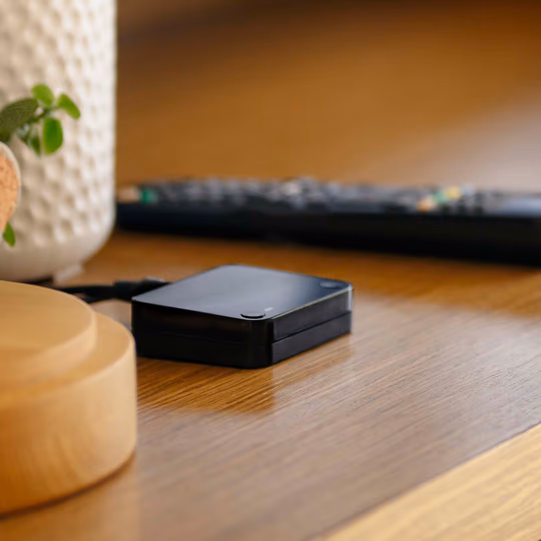 Boîtier noir compact avec câble connecté posé sur une surface en bois avec une télécommande floue en arrière-plan.