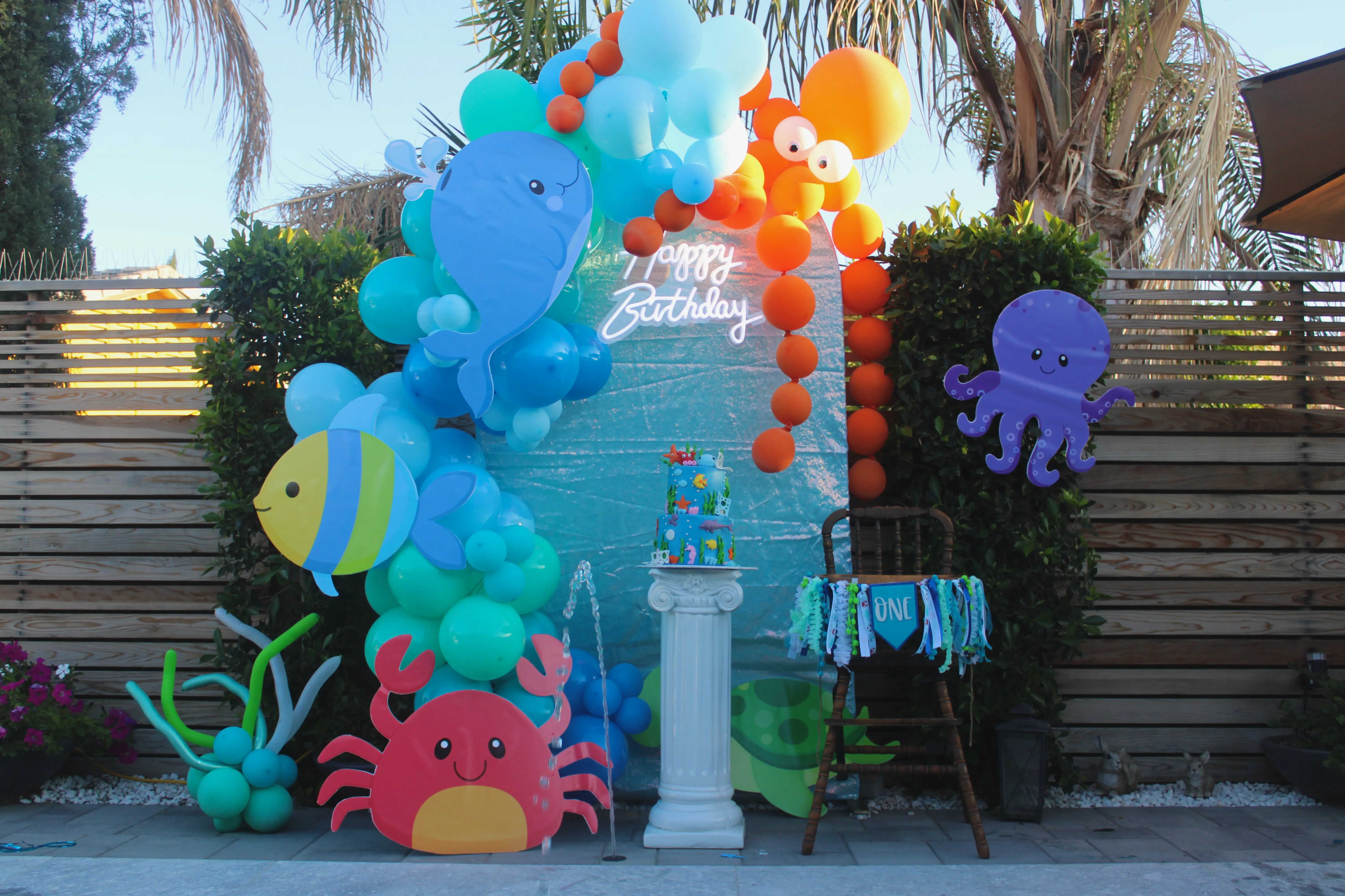 Under the sea themed first birthday party with a balloon backdrop, high chair, and cake pedestal decorated in ocean colors.