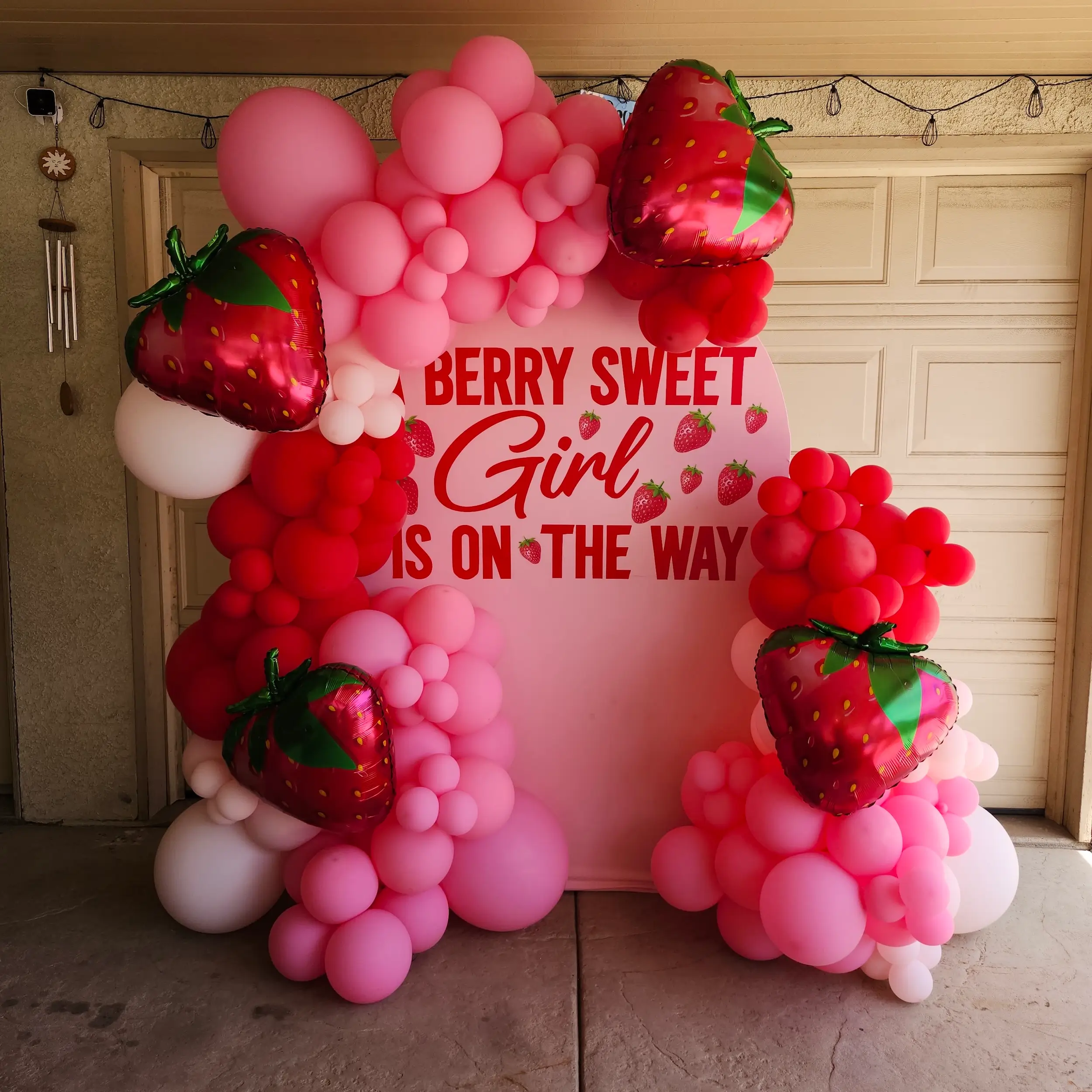 A pink and red balloon arch with strawberries and a sign reading "Berry Sweet Girl is on the Way" for a baby shower backdrop.