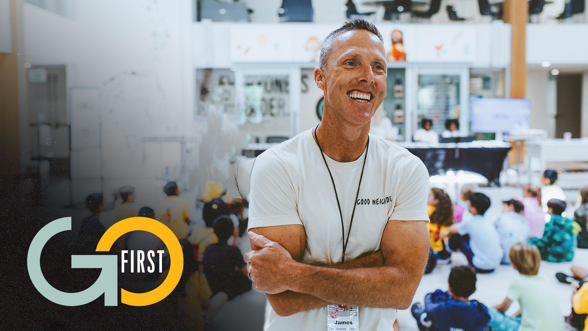 Smiling man wearing a white 'Good Neighbor' shirt and a name tag that says James, standing with arms crossed in front of children seated on the floor in a church lobby event space.