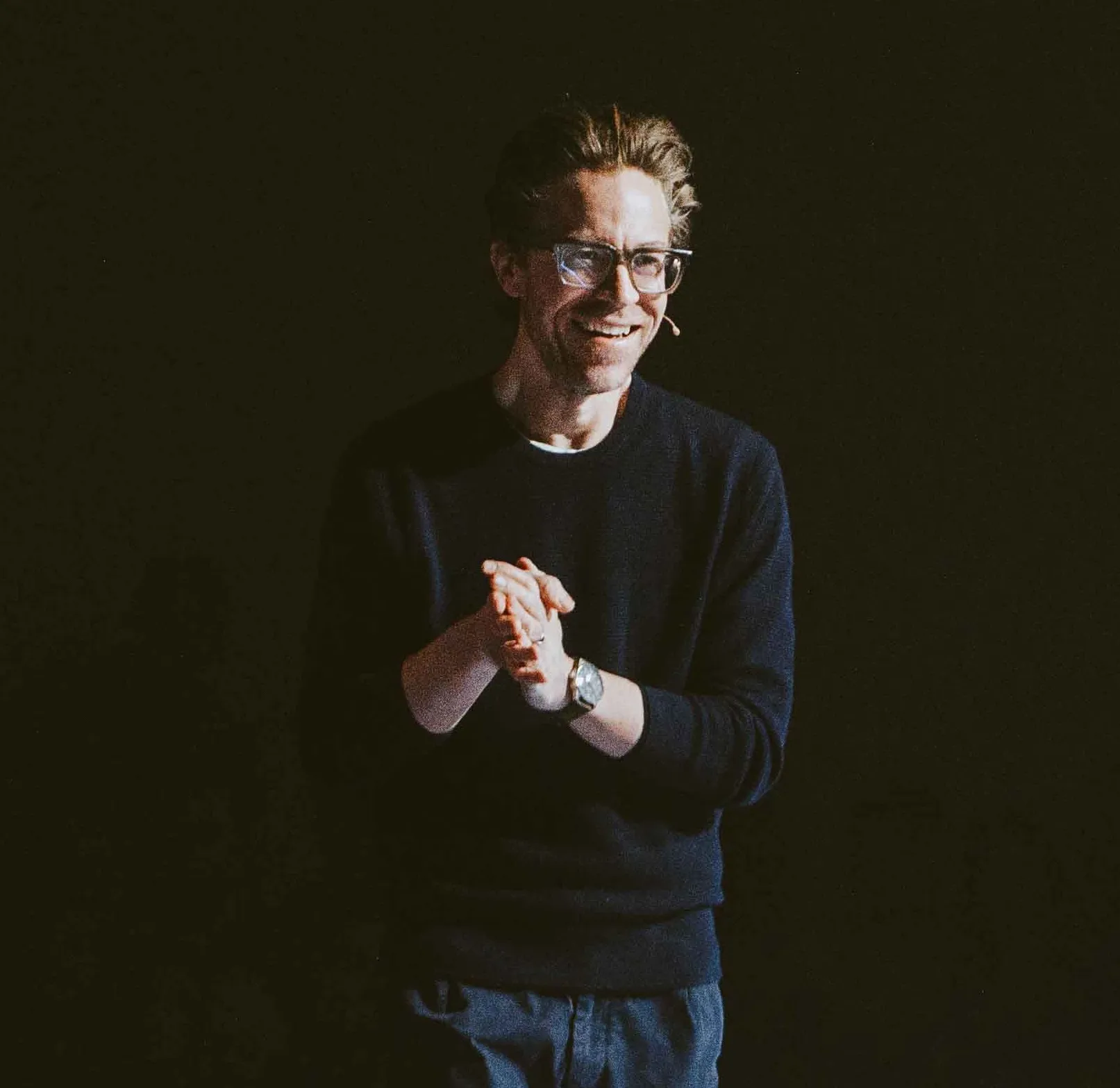 Man wearing glasses and a headset microphone, smiling and clasping hands in front of a dark background.