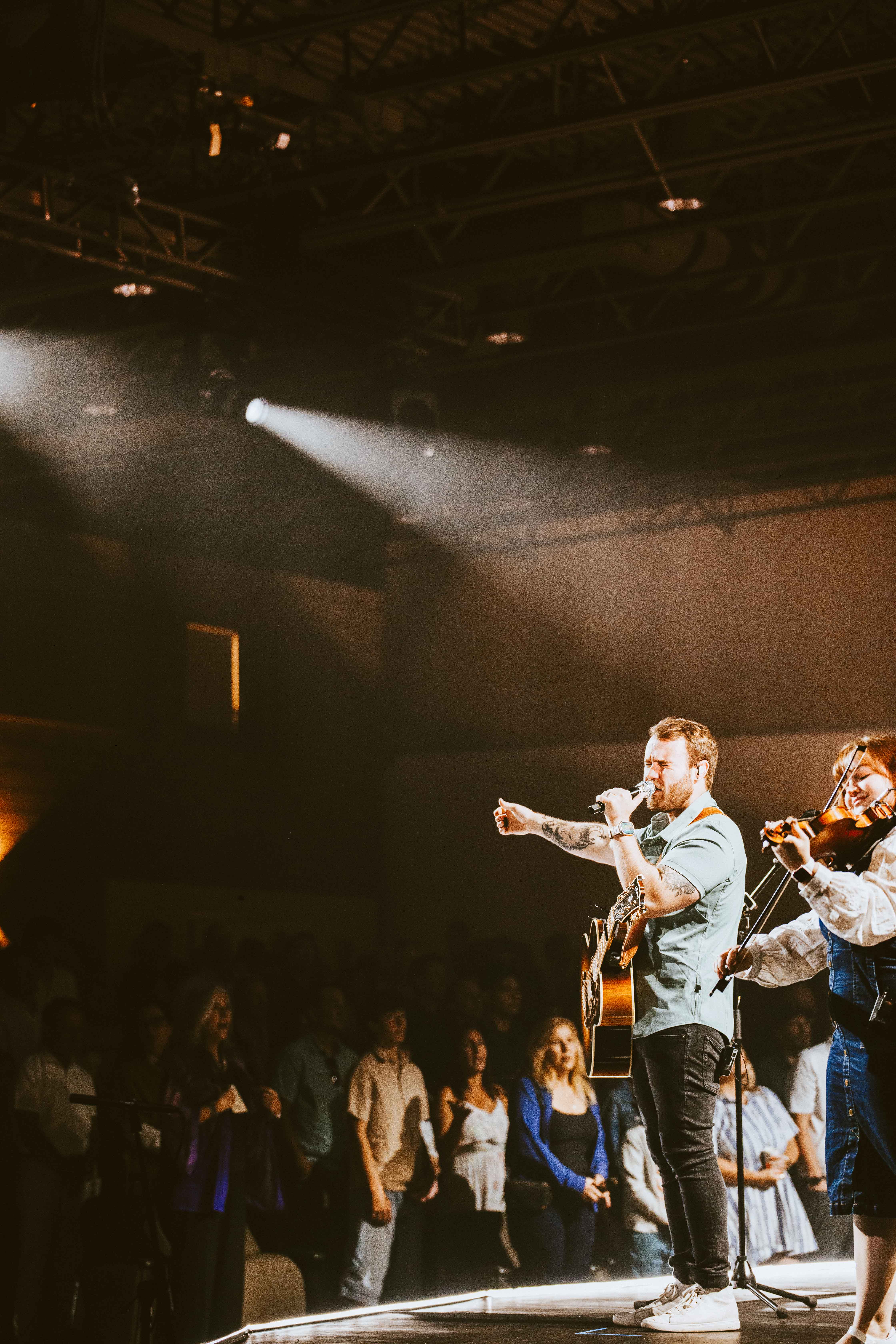 Male vocalist playing guitar and singing on stage under a spotlight, accompanied by a female violinist, with a congregation in the background.