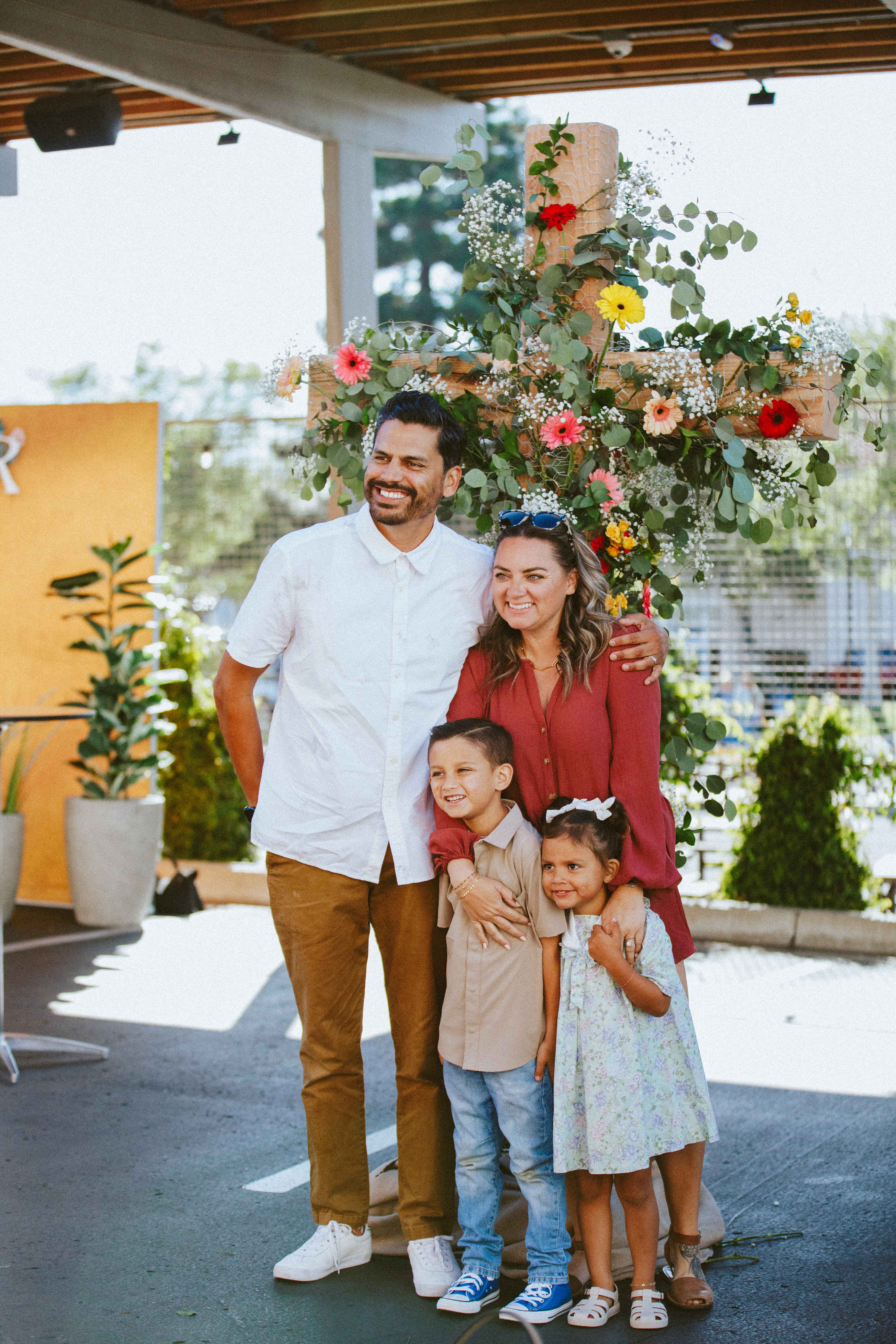 Family of four smiling and posing in front of a large wooden cross decorated with flowers and greenery.