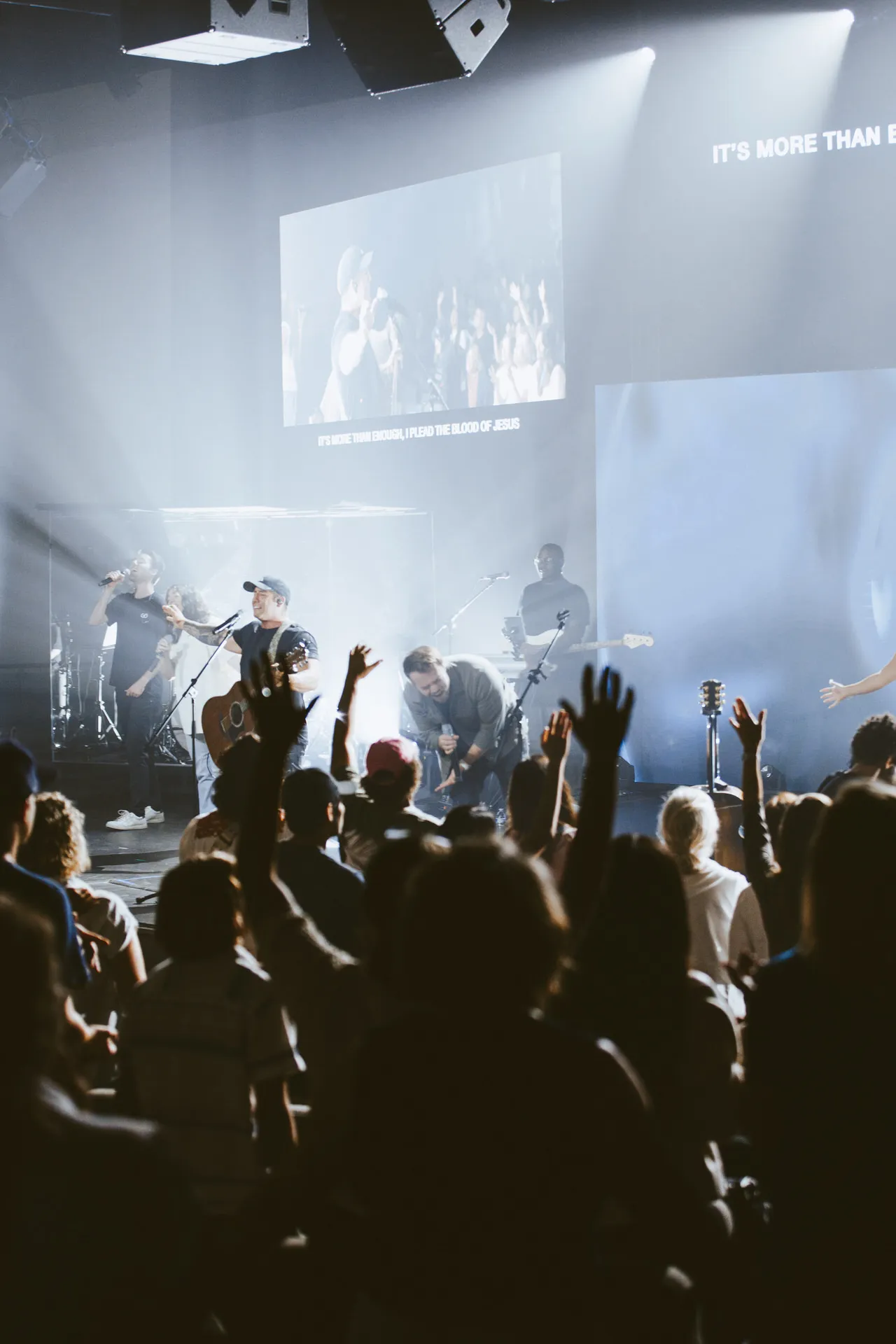 Musicians worshiping on stage with raised hands from an engaged congregation in a dimly lit sanctuary setting.