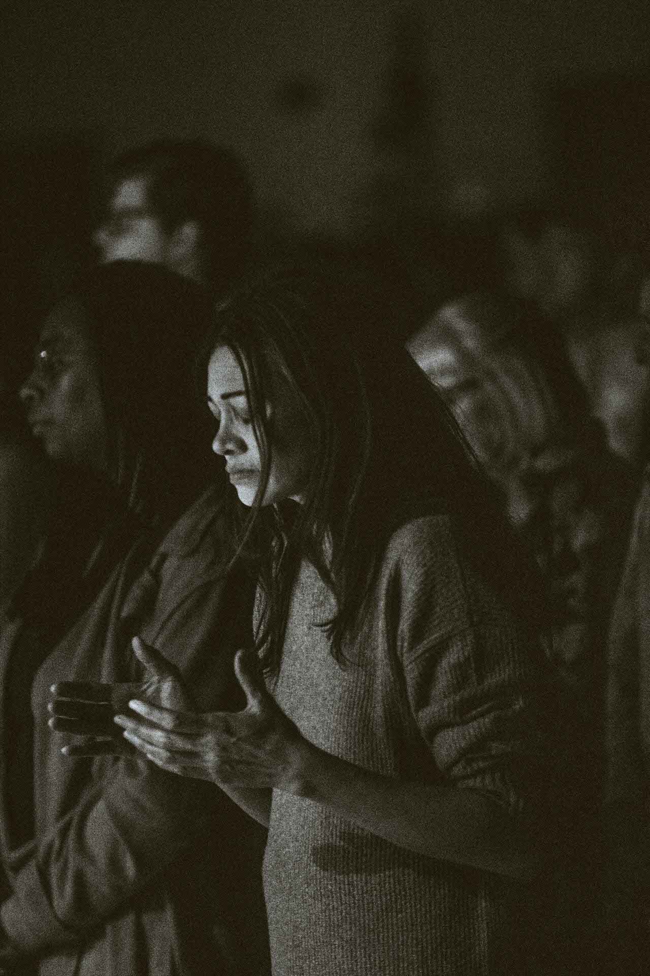 Woman with closed eyes and hands raised in a dimly lit crowd, in deep reflection during worship.