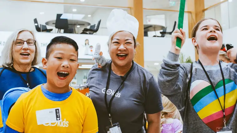 Group of smiling children and adults, including a woman wearing a chef hat, enjoying an indoor activity.