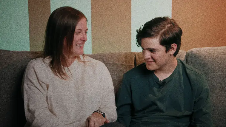A woman and a teenage boy sitting on a gray couch, smiling and looking at each other against a striped beige and light blue background.