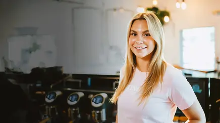 Smiling blonde woman in a light pink shirt standing in a dimly lit indoor space.