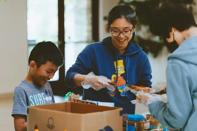 Three people, including a smiling woman in glasses and a Bart Simpson hoodie, packing food into a box.