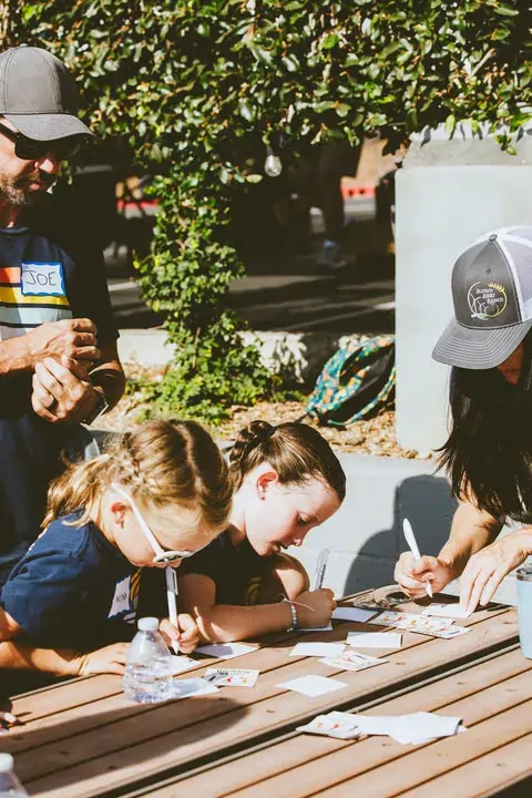Two young girls and an adult woman sitting at an outdoor wooden table writing or drawing on paper with a man standing nearby watching them.