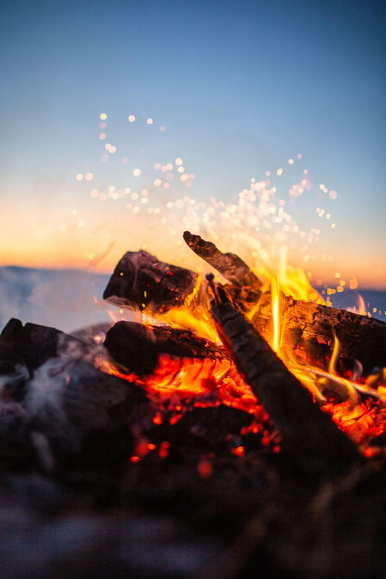 A close-up image of a crackling campfire with glowing embers and sparks against a fading sunset sky