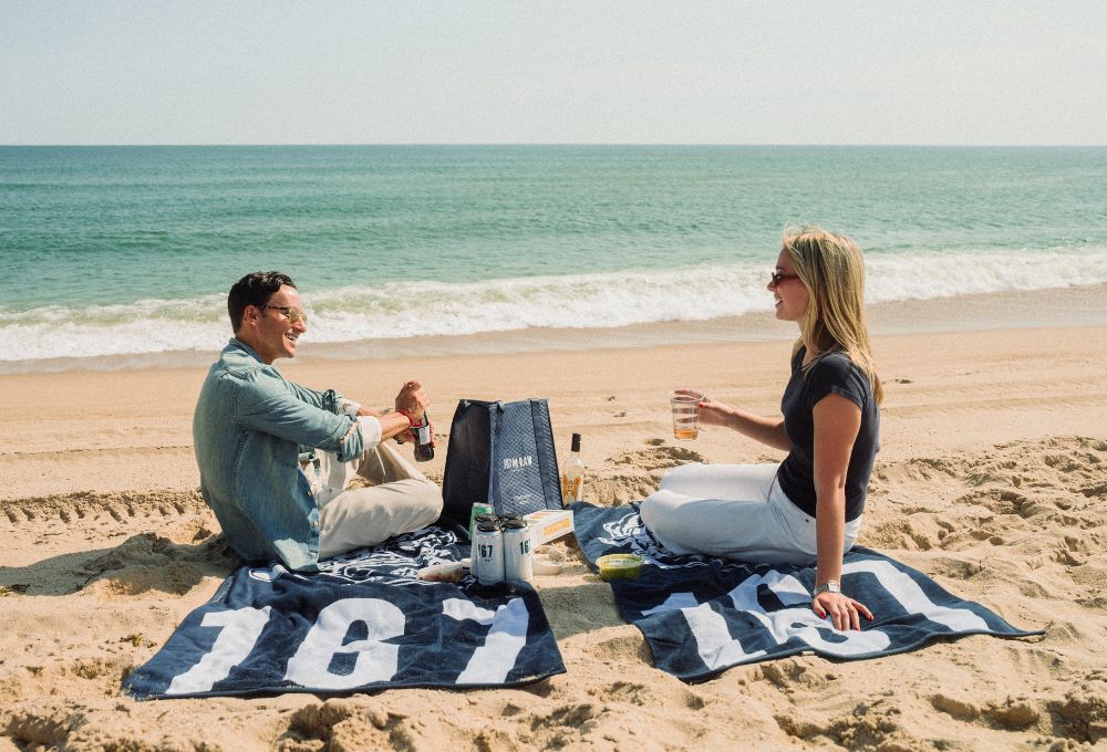Two people having drinks and snacks on the beach with 167 Raw branded towels