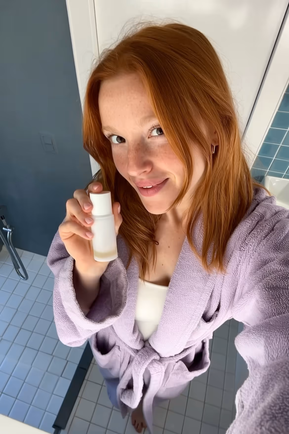 Smiling red-haired woman in a lavender bathrobe holding a small white bottle in a bathroom.