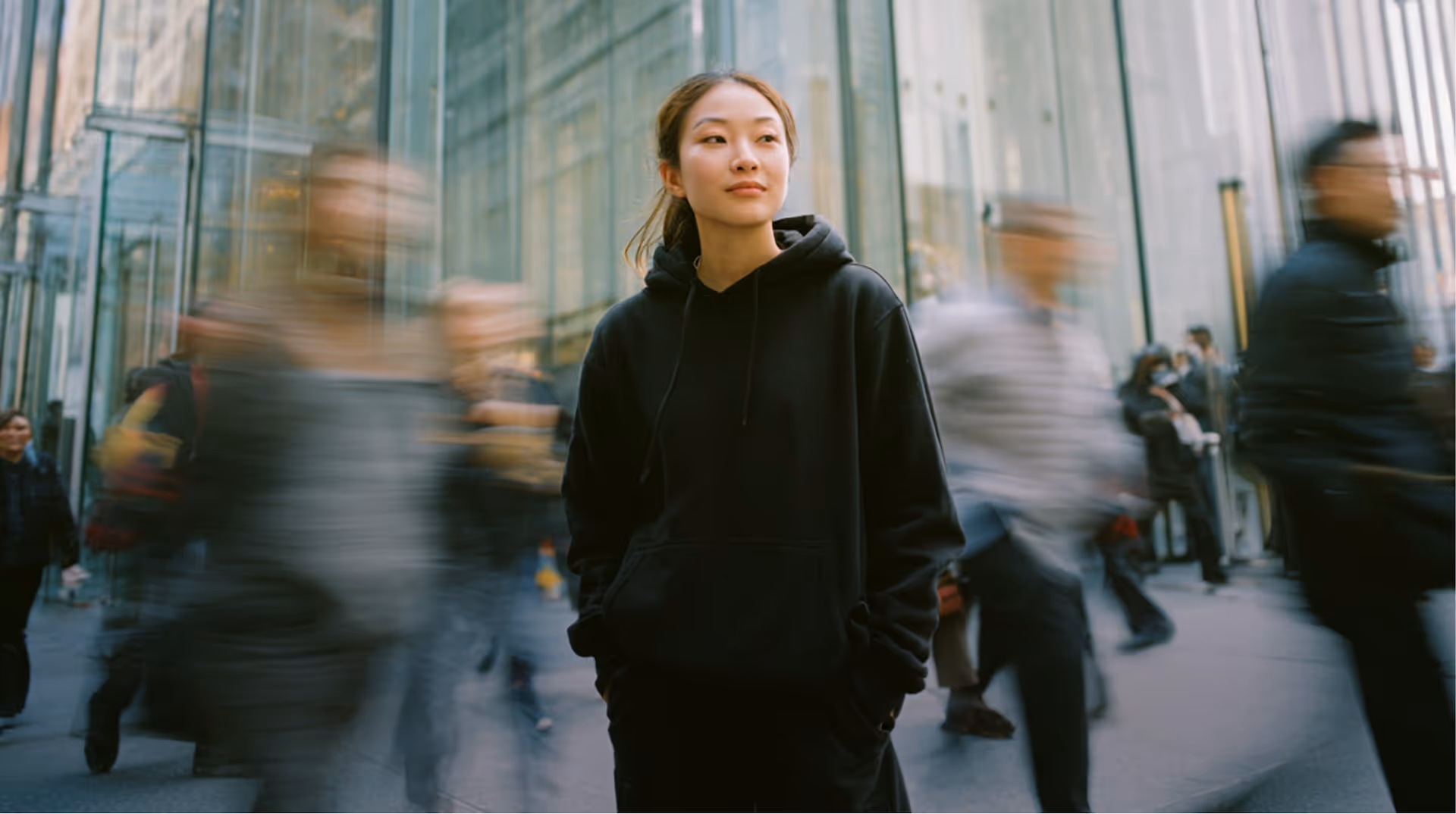 Young woman in a black hoodie standing still among blurred people walking in a busy urban setting.