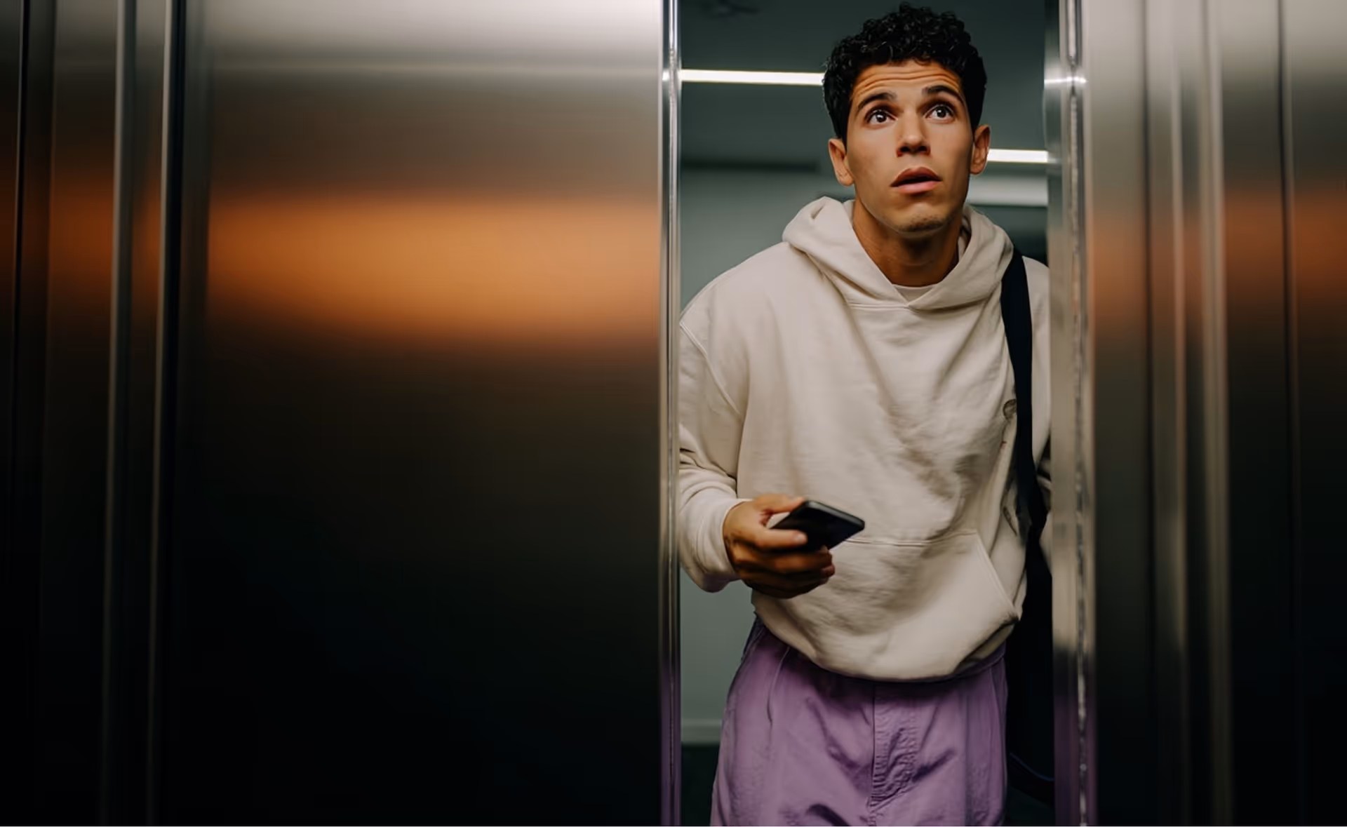 Young man in a hoodie holding a phone, looking up as elevator doors open.