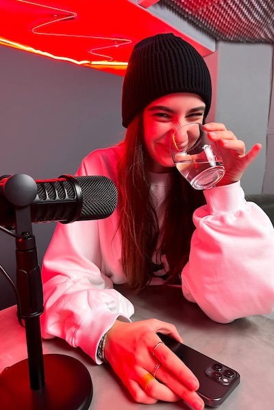 Young woman in a black beanie and white sweatshirt smiling while holding a glass of water near a microphone and smartphone on a table illuminated by red lighting.
