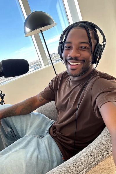 Smiling man with braided hair wearing headphones and a brown shirt, sitting on a chair near a window with a city view.