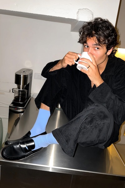 Young man with curly hair wearing black corduroy outfit and light blue socks sitting on a metal kitchen counter, drinking from a white mug.
