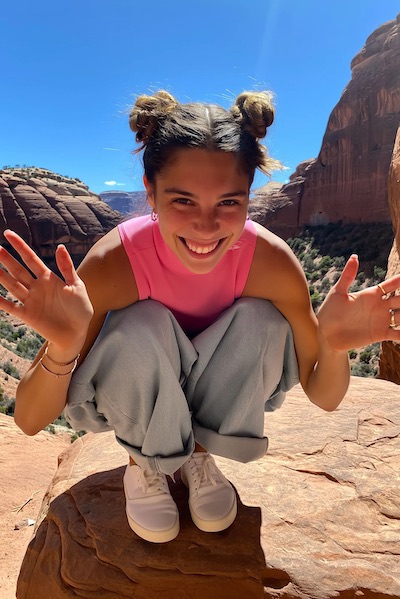 Smiling young woman with hair in buns crouching on a rock in a desert landscape with red cliffs and clear blue sky.