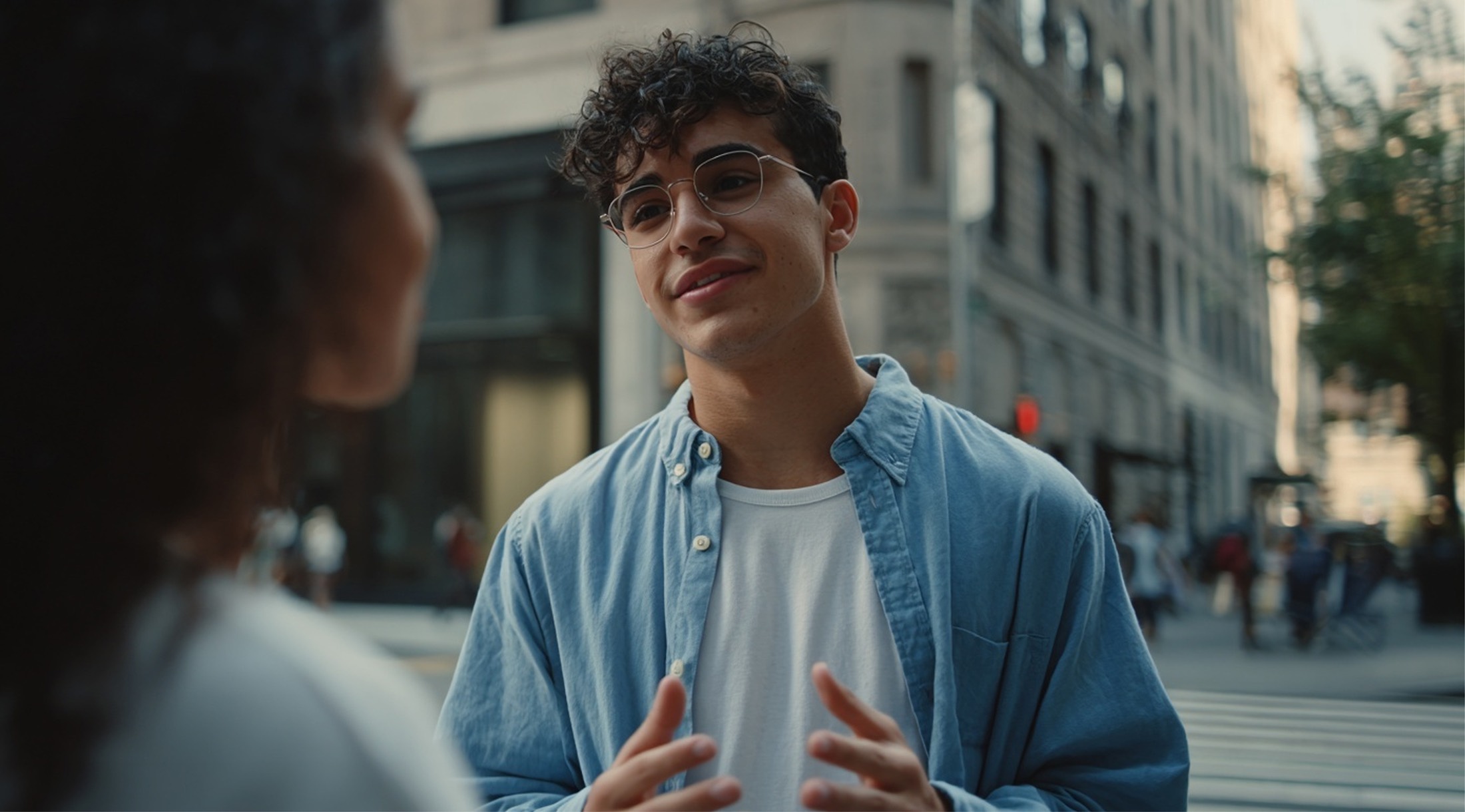 Young man wearing glasses and a blue shirt talking to a woman on a city street.