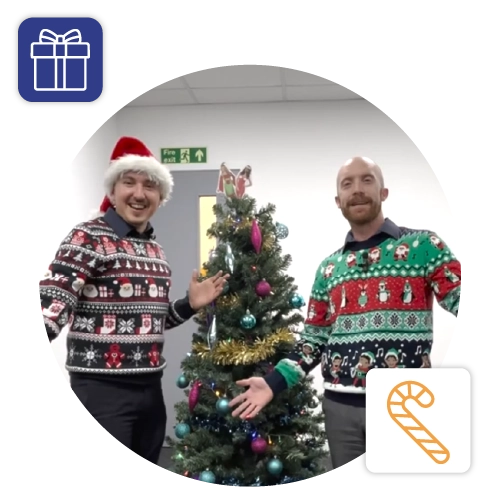 Two men in festive jumpers in front of a Christmas tree.
