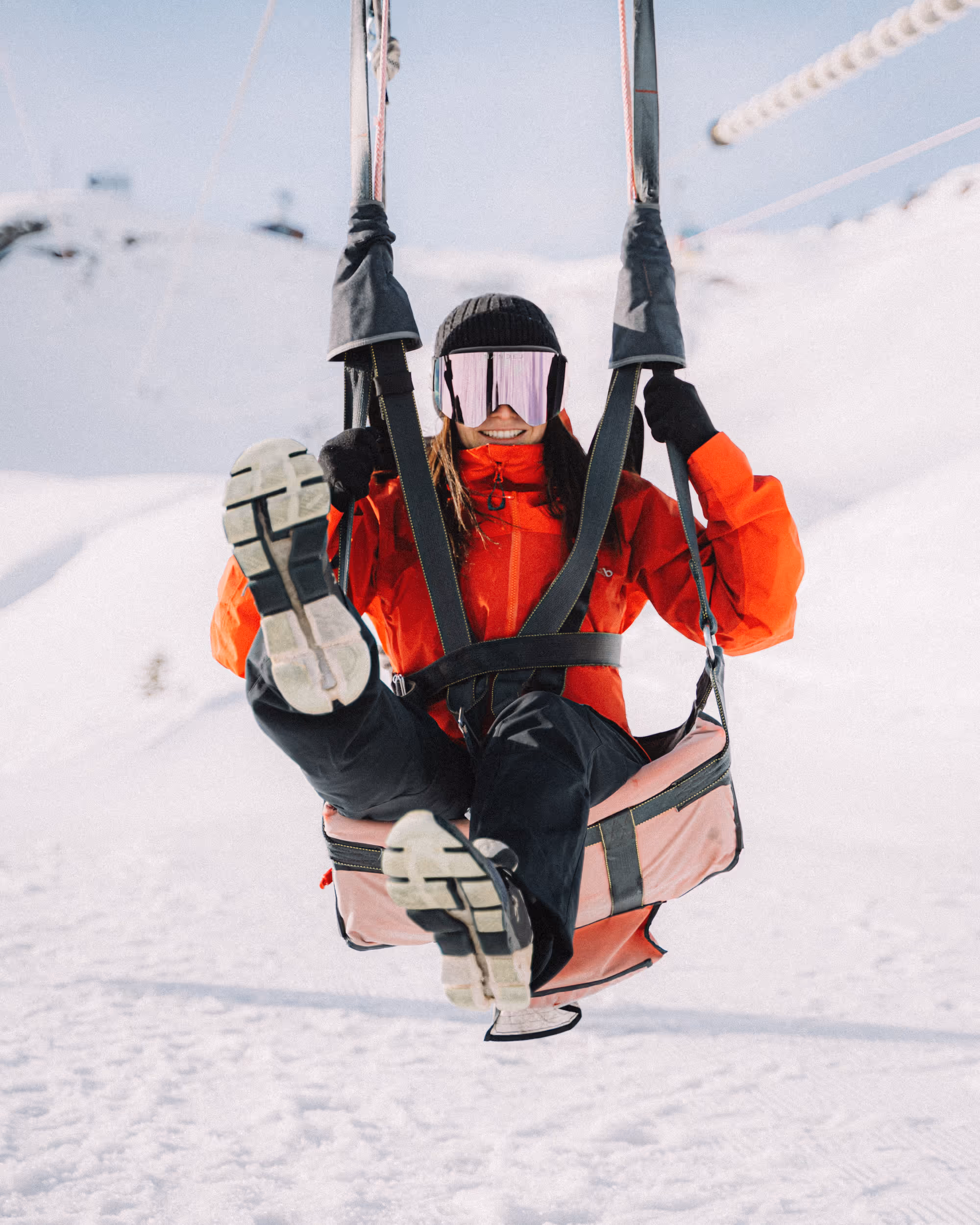 Smiling person in red jacket and ski goggles sitting on a swing in a snowy landscape.