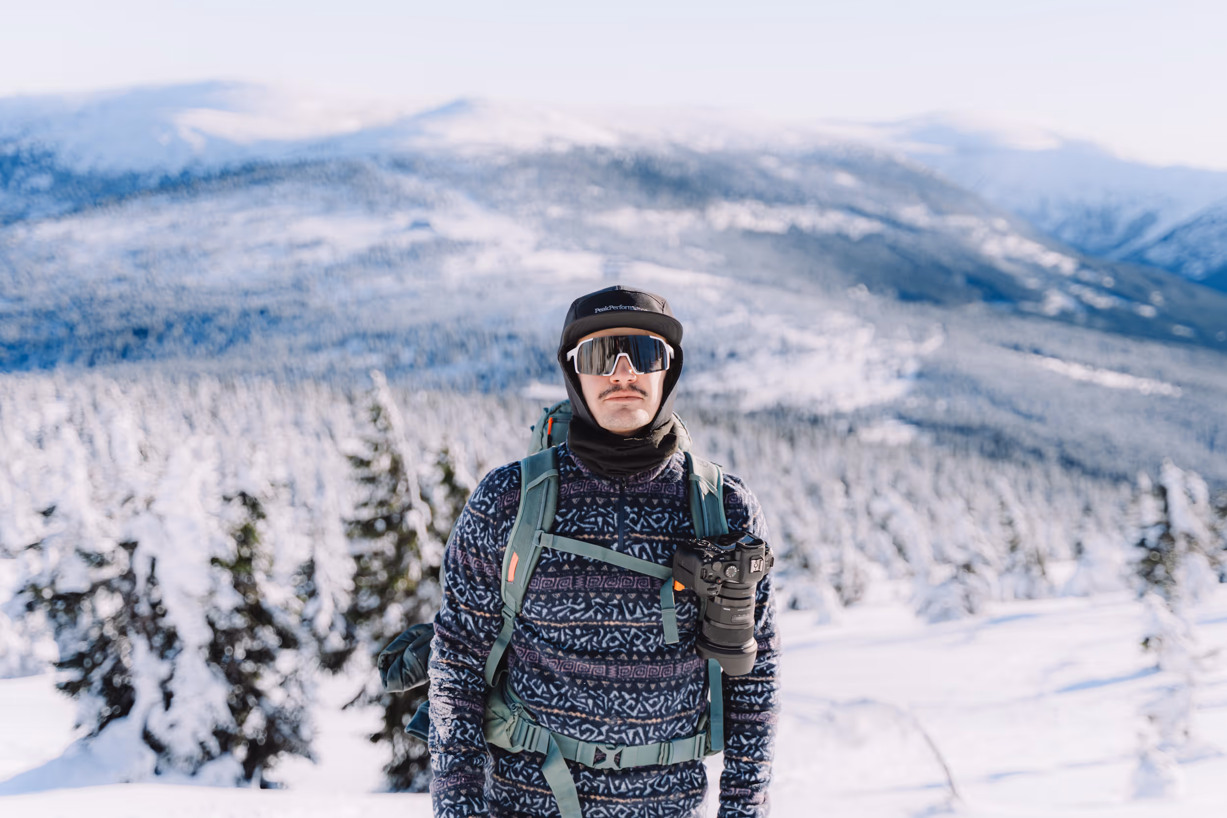 Man wearing sunglasses, a patterned sweater, and a black cap with a green backpack standing in a snowy mountain landscape.