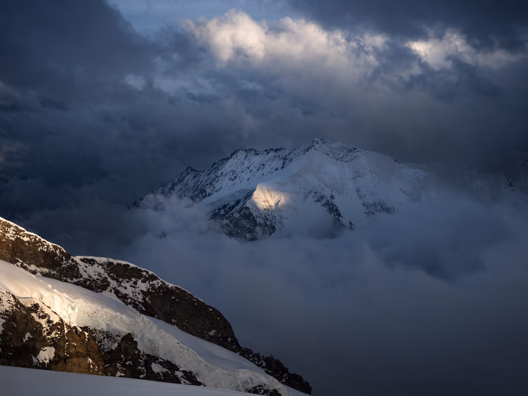 Snow-covered mountain peaks partially obscured by clouds with sunlight breaking through a dark cloudy sky.