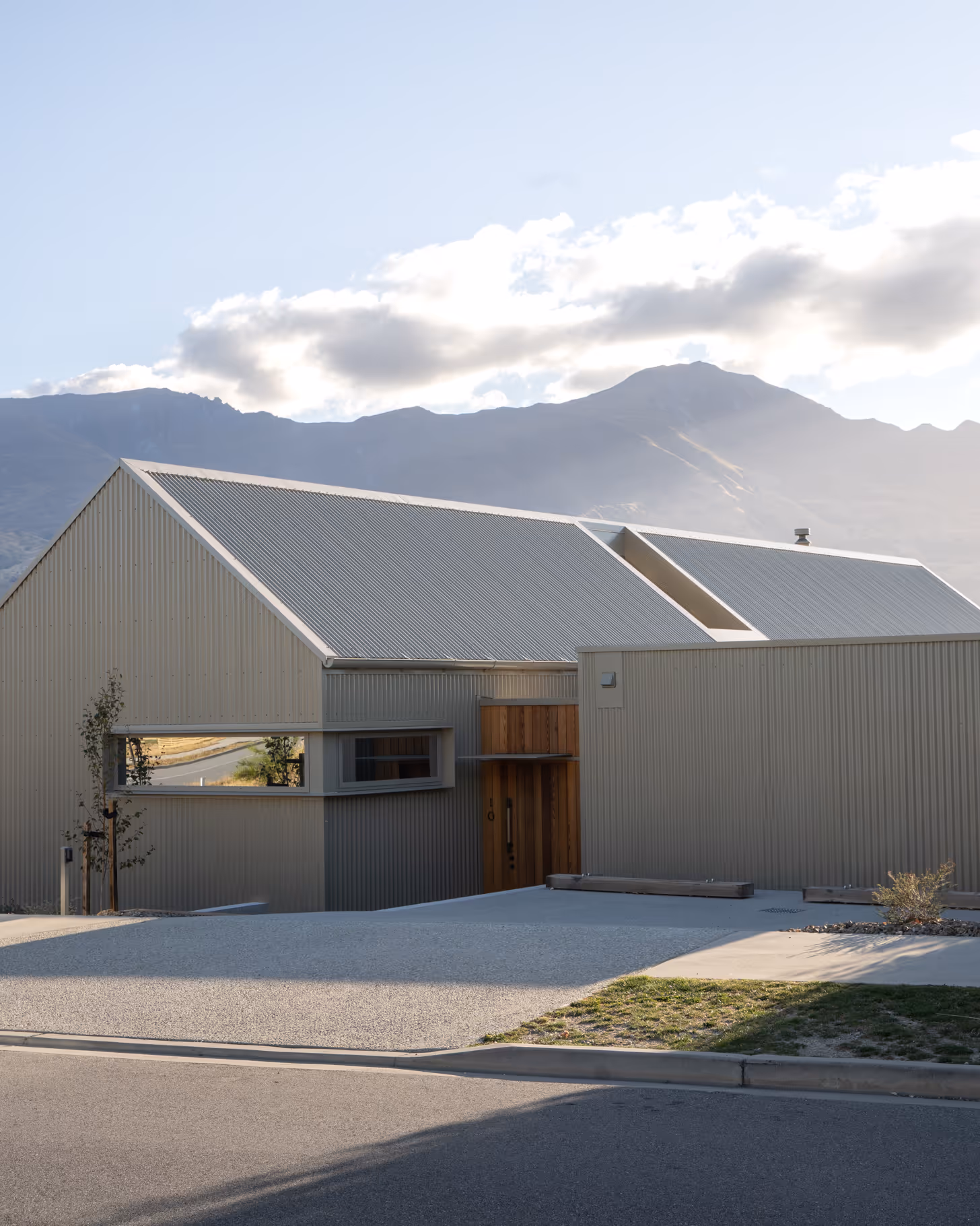 Modern house with a corrugated metal roof and siding, light beige color, wooden entrance door, set against a mountainous backdrop with partly cloudy sky.