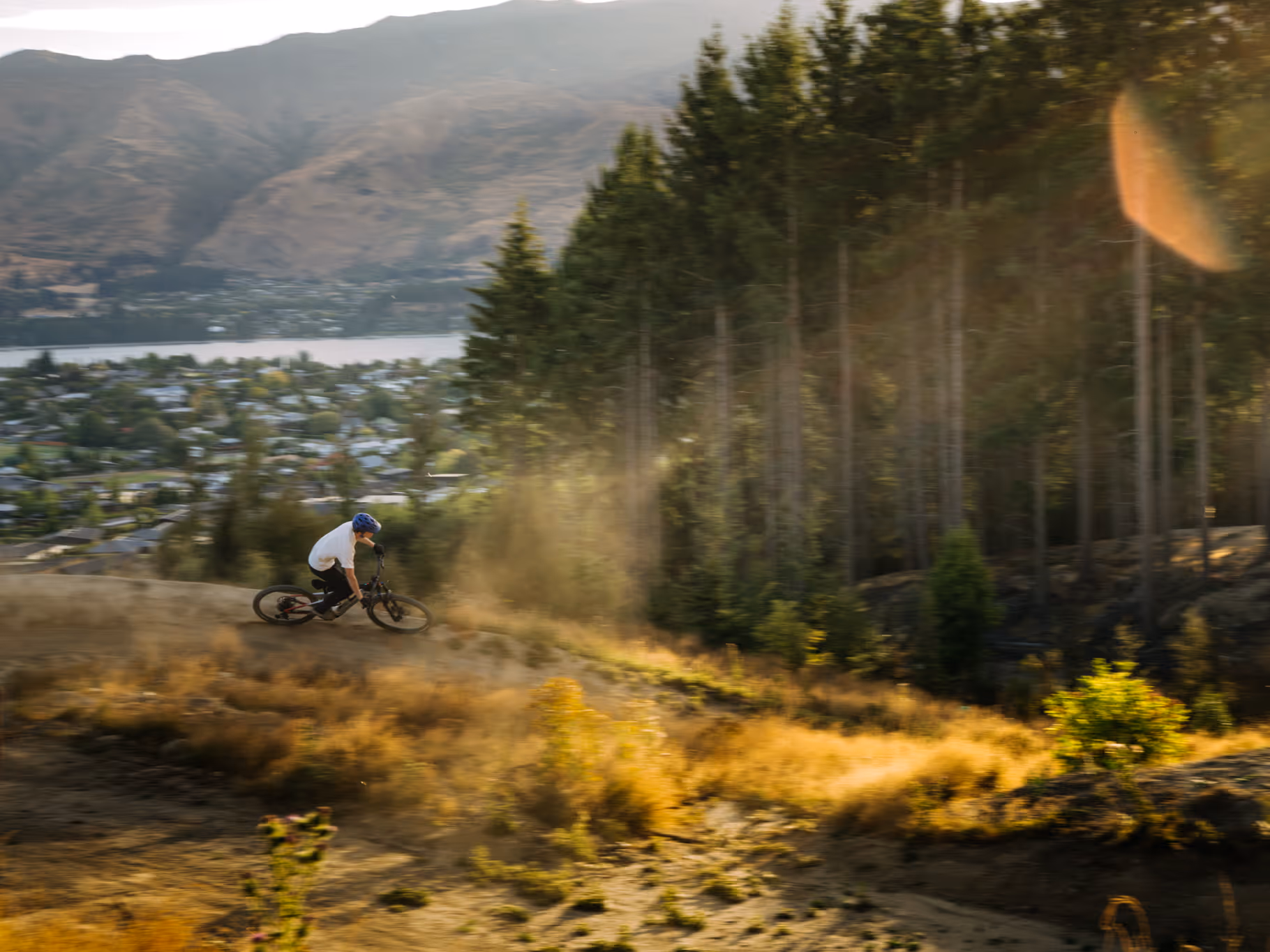 Person wearing a blue helmet rides a mountain bike downhill on a dirt trail with trees and a town visible in the background.