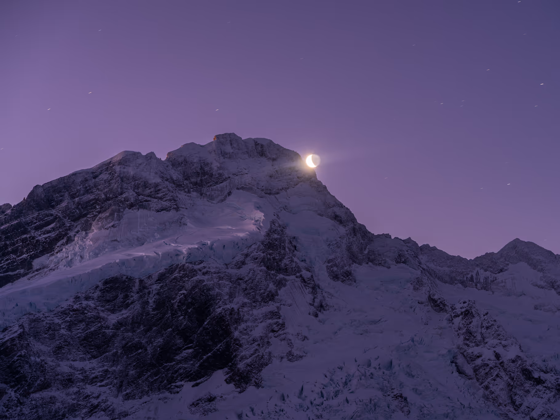 Snow-covered mountain peak at dusk with a crescent moon glowing right above the summit against a purple sky.