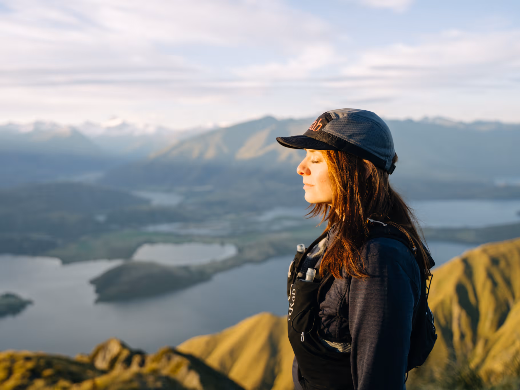 Woman in outdoor gear and cap standing with eyes closed overlooking a lake and mountain landscape.